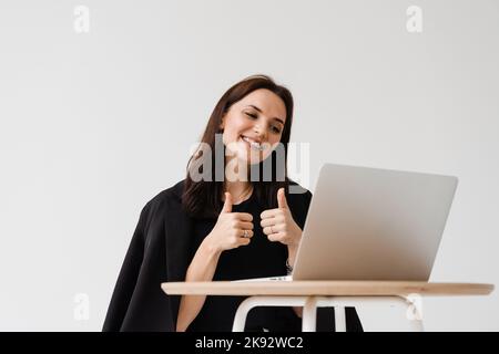 Fröhliche Frau, die ihren Kollegen und Freunden beim Videotreffen den Daumen hoch zeigt. Happy Girl mit Arbeiten auf Laptop und freut sich über die Umsetzung Stockfoto