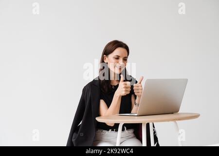 Fröhliche Frau, die ihren Kollegen und Freunden beim Videotreffen den Daumen hoch zeigt. Happy Girl mit Arbeiten auf Laptop und freut sich über die Umsetzung Stockfoto