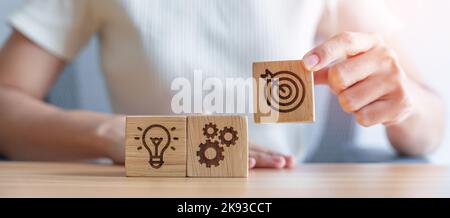 Frau Hand hält Dartscheibe über Gear und Lightbulb Icon Block. Business-Planungsprozess, Ziel, Strategie, Ziel, Mission, Aktion, Ziel, Teamw Stockfoto