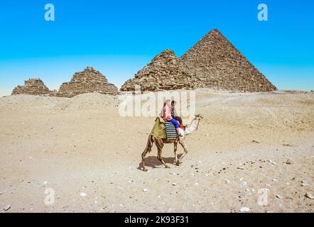 GIZA, ÄGYPTEN - 31. DEZ 1990: Touristen reiten auf dem Kamel in Gizeh in der Nähe von drei Pyramiden, die von der kleinsten, die Pyramiden der Königin und die größte, Stockfoto