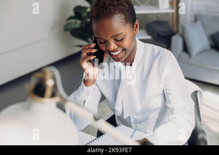 Eine hübsche afroamerikanische Frau, die am Schreibtisch telefoniert, während sie am Laptop arbeitet. Unterstützung des Kunden in einem behindertengerechten Büro Stockfoto