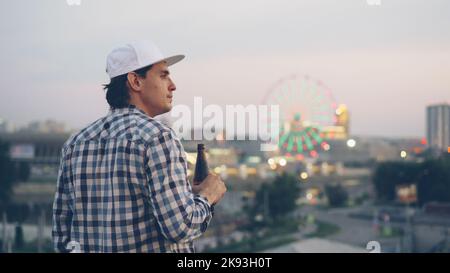 Trauriger junger Mann trinkt Bier und genießt eine wunderschöne Aussicht auf die moderne Stadt, die allein auf dem Dach mit einer Flasche steht. Junge Menschen, Stadtlandschaft und Getränkekonzept. Stockfoto