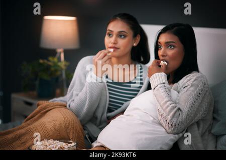 Das machen wir am Wochenende. Zwei junge Frauen essen Popcorn, während sie nachts auf einem Bett sitzen. Stockfoto