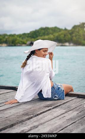 Mein Lieblingsurlaubsort ist die Nähe zum Meer. Eine attraktive junge Frau, die am Rande einer Promenade sitzt und im Urlaub eine Brise genießt. Stockfoto
