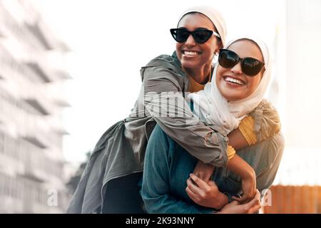 Beste Freunde wie diese sind schwer zu finden. Eine attraktive junge Frau mit Sonnenbrille und Kopftuch, während sie ihrer Freundin einen Huckepack schenkt Stockfoto