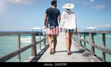 Was für ein schöner Tag, um mit Ihnen zu erkunden. Rückansicht eines unverkennbaren Paares, das während eines Urlaubs eine Promenade entlang läuft und sich die Hände hält. Stockfoto