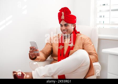 Ein Lächeln ist das Beste, was ein Bräutigam tragen kann. Ein junger Mann, der an seinem Hochzeitstag ein Smartphone benutzt. Stockfoto