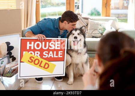 Wir waren glücklich mit unserem neuen Zuhause. Ein glückliches junges Paar und ihr Hund machten Fotos mit einem verkauften Schild in ihrem neuen Zuhause. Stockfoto