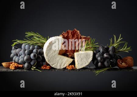 Käsekamembert mit Walnüssen, blauen Trauben und Rosmarin auf schwarzem Hintergrund. Stockfoto