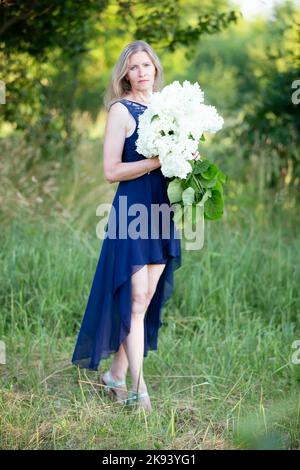 Eine schöne ältere Frau in einem blauen Kleid posiert im Garten mit einem Blumenstrauß. Stockfoto