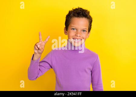 Foto-Porträt von niedlichen kleinen Jungen zeigt V-Zeichen optimistisch strahlend Lächeln tragen trendige violette Kleidungsstück auf gelbem Hintergrund isoliert Stockfoto