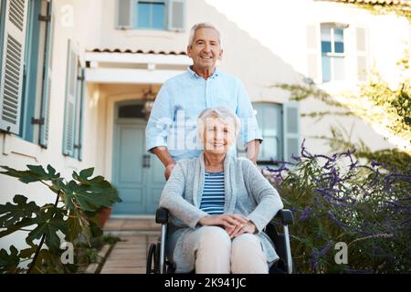 Wir steckten zusammen durch dick und dünn. Porträt einer fröhlichen älteren Frau, die im Rollstuhl sitzt und sich mit ihrem Mann zu Hause in ihrem Hinterhof entspannt. Stockfoto
