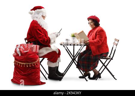 Weihnachtsmann mit Smartphone und einer älteren Frau, die an einem Tisch sitzt und auf weißem Hintergrund ein Buch liest Stockfoto
