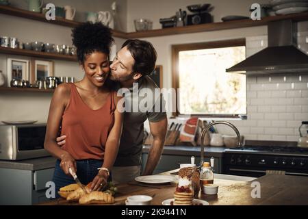 Ich habe Sie lieber zum Frühstück. Ein liebevoller Mann mittleren Alters küsst seine Frau auf die Wange, während sie das Frühstück in ihrer Küche zu Hause zubereitet. Stockfoto