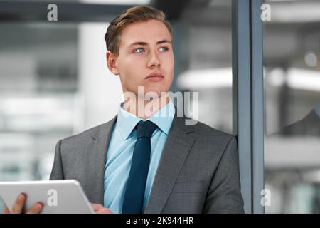 Ich denke an meinen nächsten Geschäftsumzug. Ein hübscher junger Geschäftsmann, der allein in seinem Büro steht und besinnlich aussieht, während er ein Tablet benutzt. Stockfoto