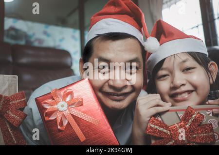 Asiatische Familie, bestehend aus Vater und Tochter tragen Weihnachtsmützen und schenken einander während des Festivals Geschenke. „Weihnachtstag“ und ein glückliches neues Jahr Stockfoto