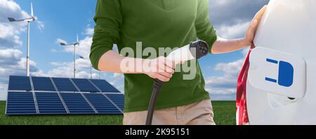 Nahaufnahme eines Menschen mit Ladekabel für Elektroautos auf dem Hintergrund von Solarmodulen und Windkraftanlagen Stockfoto