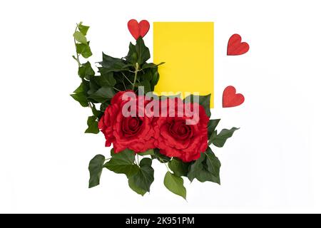 Rote Rosen auf weißem Hintergrund neben einem gelben Papier und drei roten Holzherzen, Platz für Text. Stockfoto