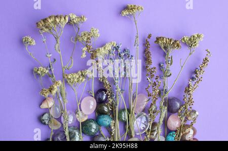 Wilde trockene Kräuter und halbedelige Kristalle aus Amethyst und Rosenquarz liegen auf dem violetten Tisch. Magische Amulette Stockfoto