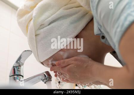 Eine junge Frau mit einem weißen Handtuch auf ihrem Kopf im Badezimmer wäscht eine Gesichtsmaske ab. Selektiver Fokus. Nahaufnahme. Hochformat Stockfoto