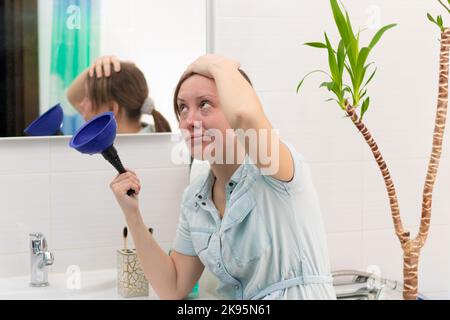 Eine junge Putzfrau in einem hellen Badezimmer mit einem Kolben, um die Verstopfung in ihren Händen zu beseitigen. An der Wand befindet sich ein Spiegel mit Spiegelung. Grüne Pflanze. Stockfoto