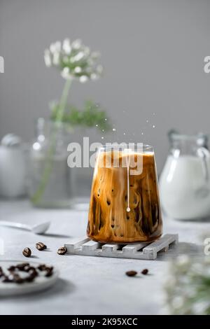 Eiskaffee in einem transparenten Glas mit Eis und Milch auf grauem Hintergrund. Fallende Milch tropft in ein Glas Kaffee. Stockfoto