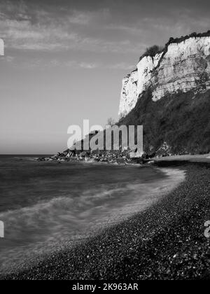 Lange Exposition, umgewandelt in Schwarz-Weiß, von Wellen, die bei Sonnenaufgang am Kiesstrand vor Kreidefelsen schwappen. Die Jurassic Küste, rühmen sich der meisten Stockfoto