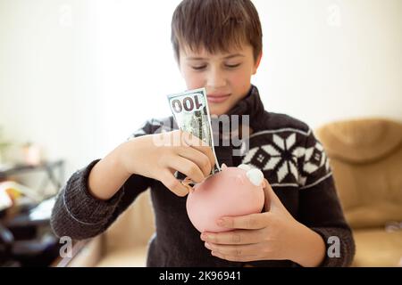 Ein Finger-Teenager legt Dollars in ein pinkfarbenes Sparschwein, das Kind lernt Finanzkompetenz. Finanzielle Einsparungen bei Jugendlichen. Ein Junge mit einem Bündel Stockfoto