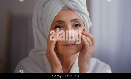 Nahaufnahme Porträt glücklich Senior Dame mit Anti Falten Haut Hydrogel Kollagen Flecken unter den Augen lächeln Kamera alte 60s kaukasische Frau mit Handtuch Stockfoto