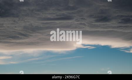 wolkiger, unheimlicher Himmel, blassweißer blauer Himmel mit verstreuten Cumulus-Wolken vor Regen. Eine grau-blaue Wolke wird von einer Windströmung bewegt, Wolken vor Regen Stockfoto