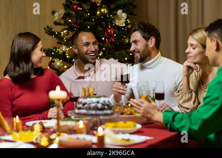 Freunde feiern Weihnachtsabend oder Silvester paty zusammen am Tisch sitzen. Schlemmen Sie zu Hause Gruppe von multi ethnischen festlichen weihnachtsessen. Glückliche fröhliche Menschen klirken Weingläser lachen Stockfoto