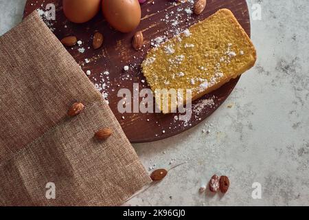 Ein rundes Holzbrett mit Backzutaten wie Mandeln, Eiern, Mehl und einem Stück hausgemachten Kuchen Stockfoto
