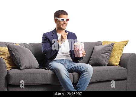 Junger Mann mit 3D Gläsern, der Popcorn isst und auf einem isolierten Sofa auf weißem Hintergrund sitzt Stockfoto