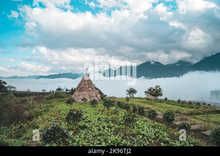 Hütten im Wigwam-Typ mit Strohdach. Strohwigwam auf einer Lichtung an einem sonnigen Frühlingstag gegen den bewölkten Himmel und die Berge. Wunderschöne Naturlandschaft. Traditionell Stockfoto