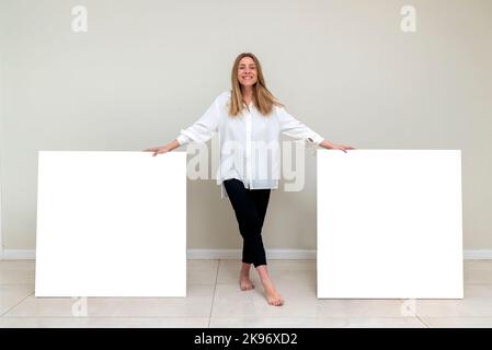 Eine junge Frau in einer weißen Bluse und schwarzen Hose posiert mit großen leeren Leinwänden mit Kopierraum Stockfoto