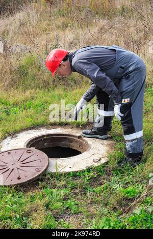 Ein männlicher, arbeitend Klempner in Overalls strahlt eine Taschenlampe in einen Wasserbrunnen. Inspektion und Reparatur von Sanitäranlagen. Stockfoto