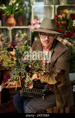 Müde oder denkende Emotionen. Attraktive kaukasische männliche Person in Brillen und Hut sitzend mit alter Schreibmaschine im Blumenladen. Dichter oder Schriftsteller Arbeitskonzept. Hochwertiges vertikales Bild Stockfoto