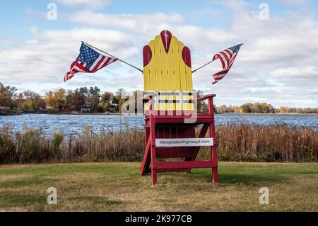 Großer roter Sessel am North Center Lake mit einer PFD darauf, weil er in den See geblasen wurde, mit zwei amerikanischen Flaggen in Center City, Minnesota, USA. Stockfoto