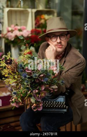 Dichter oder Schriftsteller Denken Konzept. Fröhliches kaukasisches Männerportrait in Brillen und Hut, das mit einer alten Schreibmaschine im Blumengeschäft sitzt. Hochwertige Fotos Stockfoto