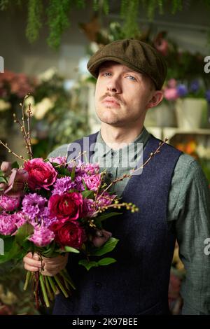 Konzept zum Muttertag oder Valentinstag. Ernste Emotion: kaukasischer Rüde in Spitzenmütze und Vintage 20s Kleidung mit einem Bouquet aus roten und violetten Blüten. Herzlichen Glückwunsch mit Blumen Stockfoto