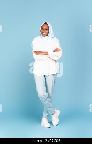 In voller Länge Porträt einer lächelnden afroamerikanischen muslimischen Frau im Kopftuch mit gefalteten Armen auf blauem Hintergrund Stockfoto
