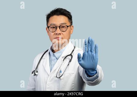 Selbstbewusster, ernsthafter chinesischer Arzt mittleren Alters in weißem Mantel, Schutzhandschuhen und Brille zeigt Stoppschild Stockfoto