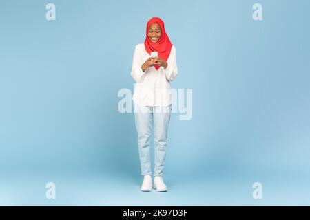 Ganzkörperaufnahme einer afroamerikanischen Frau in Hijab mit Smartphone über blauem Studiohintergrund Stockfoto