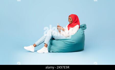Glückliche afroamerikanische muslimische Frau mit Handy und SMS online, sitzt in Sitzsack Stuhl, Panorama, freien Raum Stockfoto