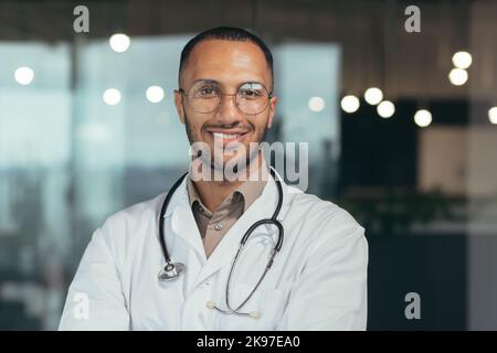 Nahaufnahme. Porträt eines jungen gutaussehenden hispanischen Arztes. In der gläsernen Halle des Krankenhauses stehend, mit Brille, weißem Mantel und Stethoskop. Er schaut lächelnd auf die Kamera Stockfoto