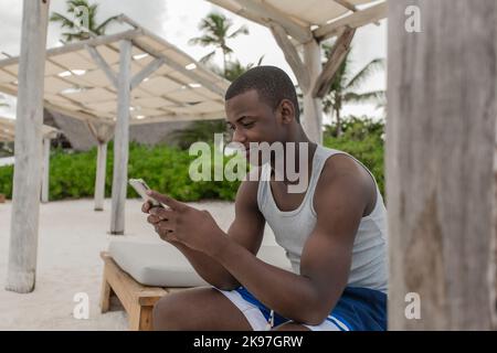 Nahaufnahme eines schwarzen Mannes im aktiven Ohr, der auf einer Holzbank sitzt und auf dem Smartphone surft, während er sich am Sandstrand ausruht Stockfoto