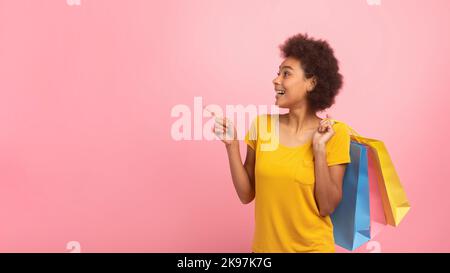 Glückliche junge Mischrasse Weibchen mit lockigem Haar mit vielen Taschen mit Käufen, zeigt Finger auf leeren Raum Stockfoto