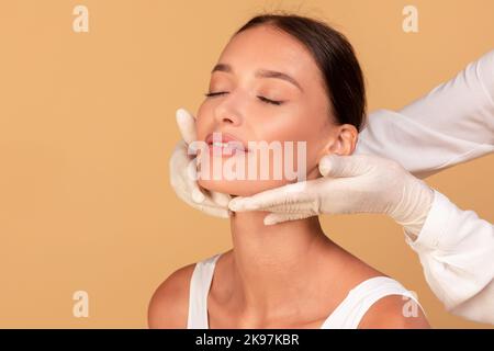 Junge Dame mit glatter Haut erhalten Inspektion des Gesichts in der medizinischen Einrichtung, Kosmetikerin in Gummihandschuhen berühren Hals Stockfoto