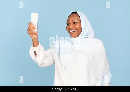 Positive afroamerikanische muslimische Frau hält Handy und macht Videoanruf oder macht Selfie auf blauem Hintergrund Stockfoto