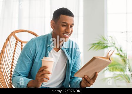 Lächelnder, nachdenklicher junger schwarzer Mann liest in lockerer Atmosphäre Buch, genießt in der Freizeit eine Tasse Getränk zum Mitnehmen, sitzt im Sessel Stockfoto